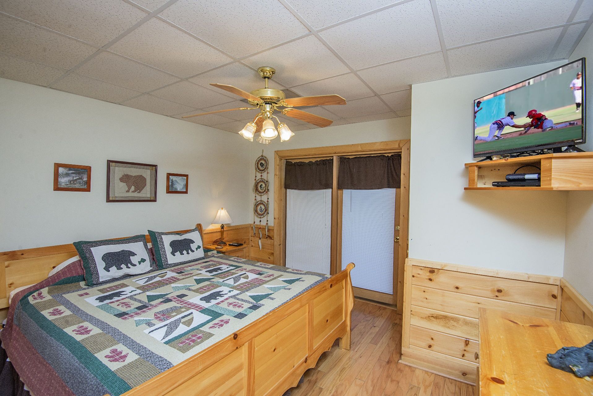A bedroom with a wooden bed, a ceiling fan, a television on a wooden shelf, and a window with blinds.