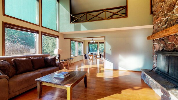 Un salon spacieux doté d’un plancher en bois, d’une cheminée en pierre et de grandes fenêtres offrant une vue sur l’extérieur.