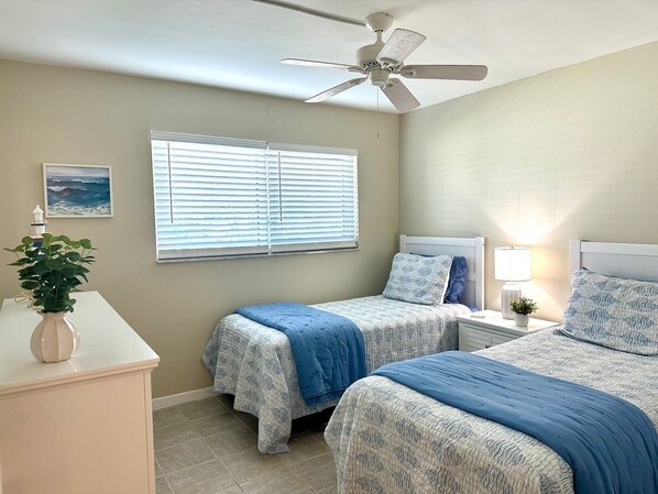 A bedroom with two beds, a ceiling fan, a window with blinds, a bedside table with a lamp, and a small plant.