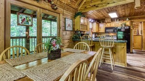 A wooden cabin interior with a dining area, kitchen, and large windows.