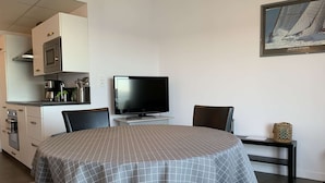 A compact hotel room with a dining table, two chairs, a flat-screen TV, a small side table, and a kitchen area with a microwave and coffee maker.