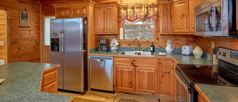 A cabin-style kitchen with wooden cabinets, a stainless steel refrigerator, a dishwasher, a sink with a window above, and a microwave.