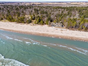 En vy över en strand, tagen från luften, med ett hus och ett skogsområde i bakgrunden.