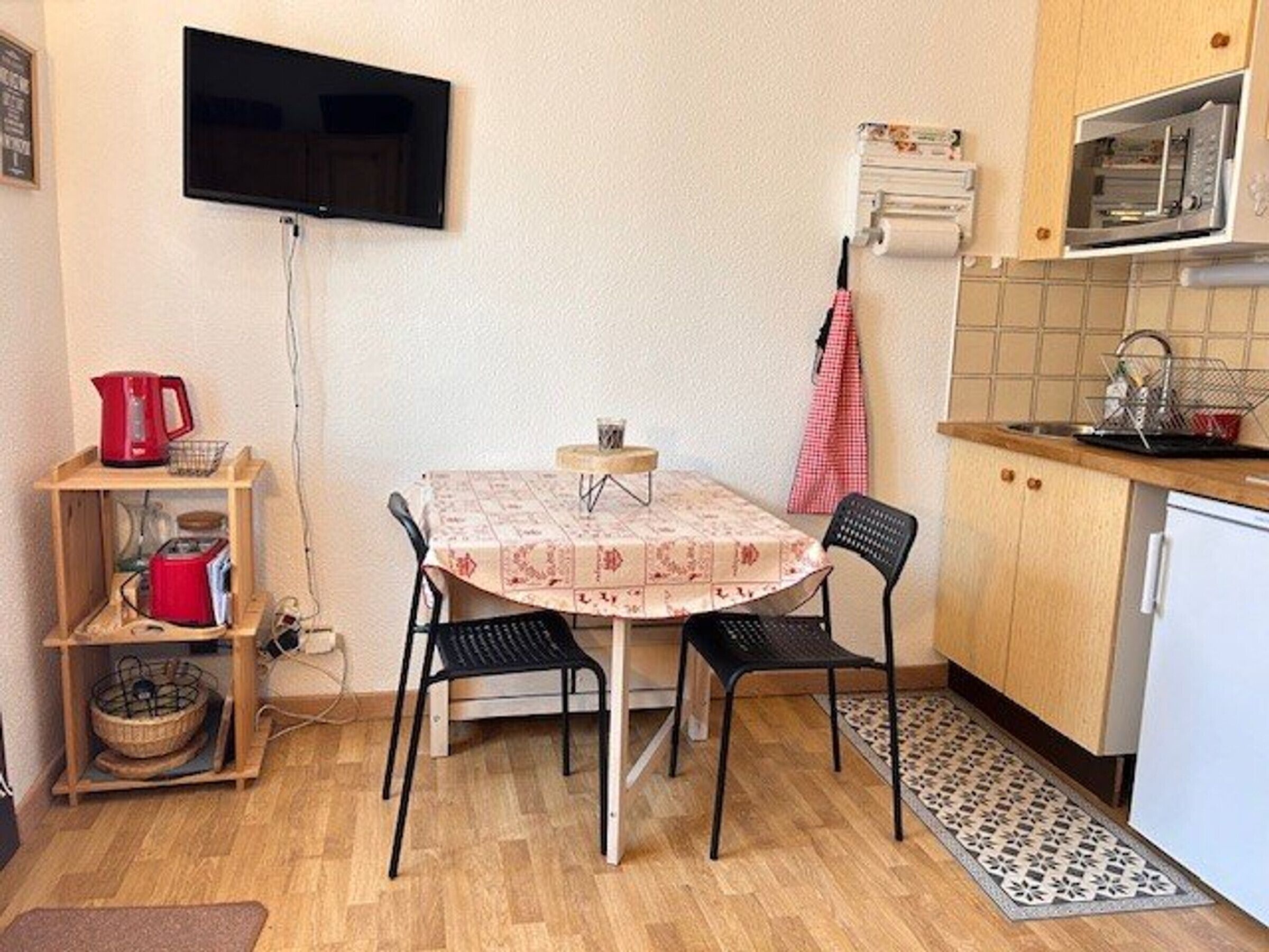 Uma cozinha compacta com mesa de jantar, cadeiras, uma TV fixada na parede, micro-ondas e uma pia pequena.