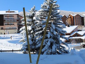 Un albero innevato di fronte a un edificio con diversi balconi e una stazione sciistica sullo sfondo.