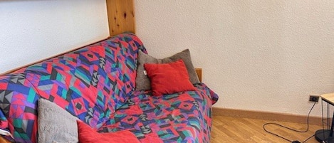 A wooden sofa with colorful patterned upholstery, a grey throw, and red pillows.