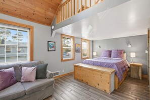 A bedroom with a large bed, a wooden bench, a gray sofa, and a window overlooking a house.
