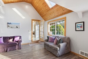 A spacious room with a high ceiling, a wooden beam structure, a grey sofa with cushions, a purple sofa, a large window with a view of greenery, and a framed picture on the wall.