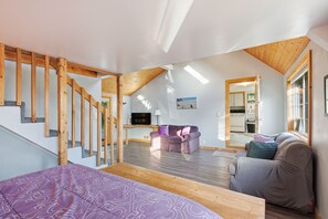 A spacious loft with a high ceiling, wooden beams, a staircase, a living area with a sofa and a TV, and a kitchen visible through an open door.
