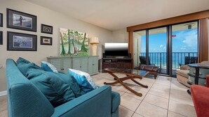 Ein Wohnzimmer mit einem türkisblauen Sofa, einem Glastisch, einem Flachbildfernseher und einem Balkon mit Meerblick.