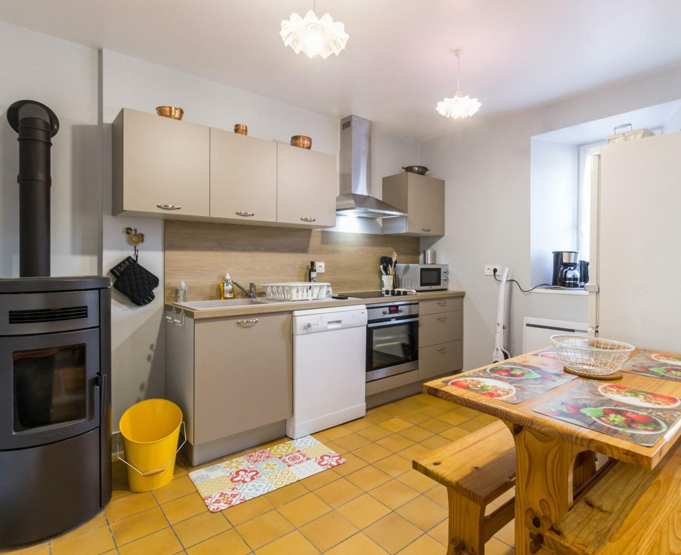 Una cocina funcional con estufa, microondas y horno, una mesa de comedor de madera con individuales, y un cubo amarillo en el suelo.