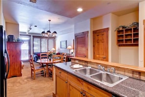A kitchen with wooden cabinets, granite countertops, and a double sink. There is a dining area with a table and chairs, and a ceiling fan. - 2109 Timberline Lodge, Trappeur's Crossing Apartment 1 (Steamboat Springs)