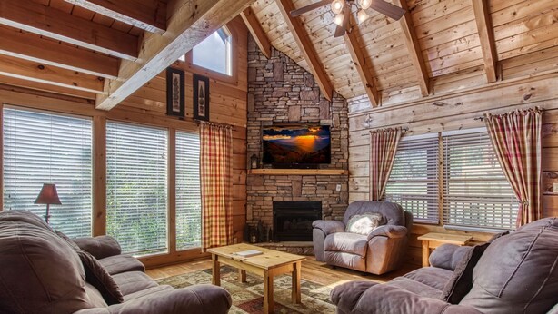 Un intérieur de chalet chaleureux avec une cheminée en pierre, des poutres en bois et de grandes fenêtres.