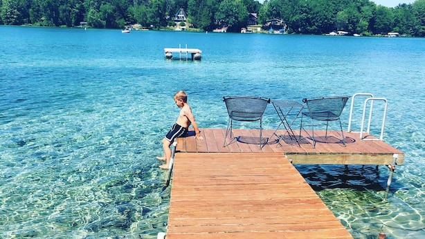 A wooden dock extending into clear blue water with a child standing at the end. There are two metal chairs and a table on the dock.