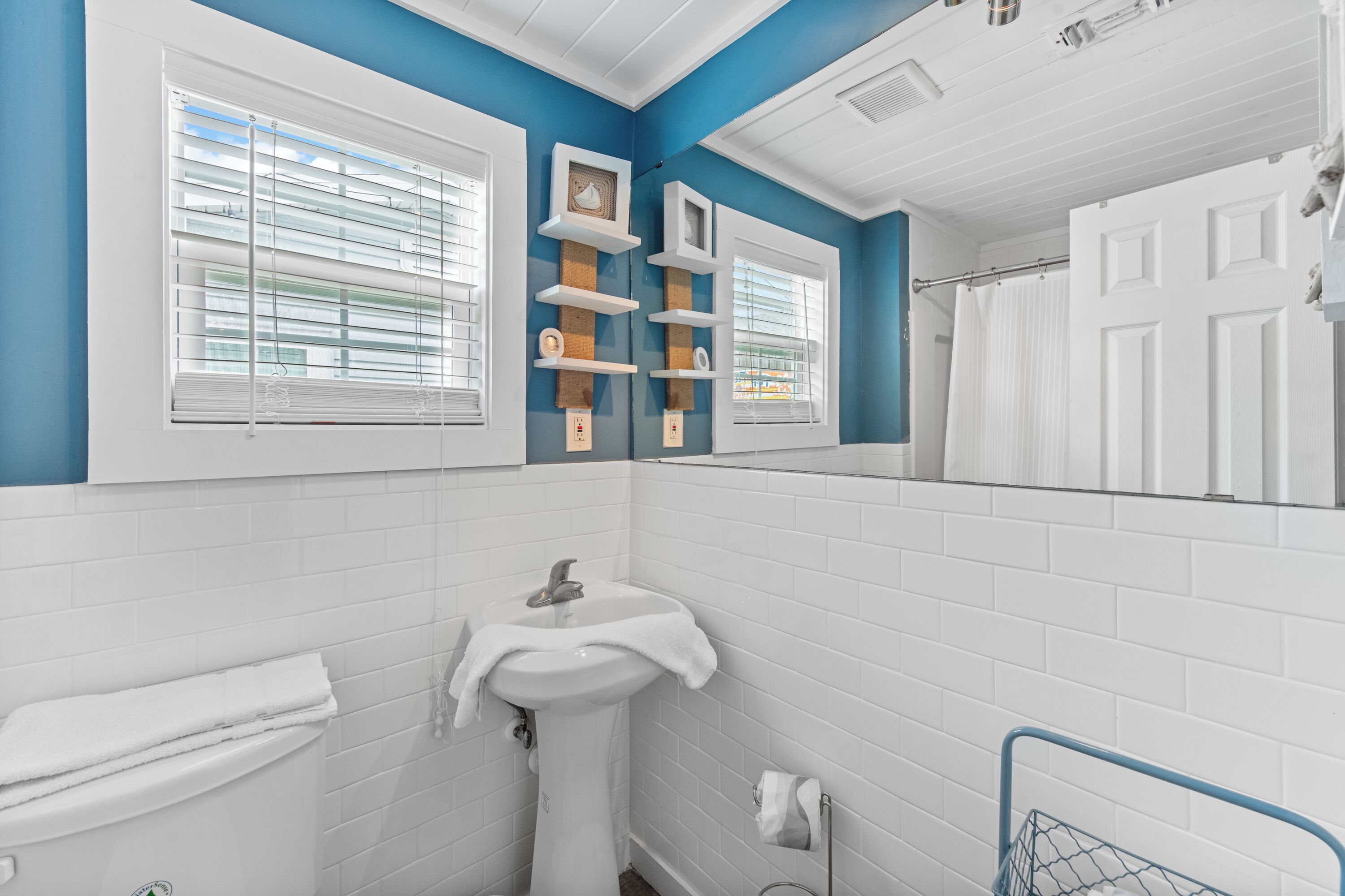 Un baño con azulejos blancos, un lavabo, un perchero para toallas y una ventana con persianas.