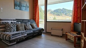 Uma sala de estar moderna com um sofá adornado com nomes de cidades, uma pintura de uma paisagem nevada, uma grande janela com vista para colinas e uma televisão sobre um suporte de madeira.