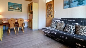 Uma sala de estar aconchegante com teto de madeira, um sofá decorado com nomes de cidades, um espaço de jantar com cadeiras de madeira e uma cozinha ao fundo.