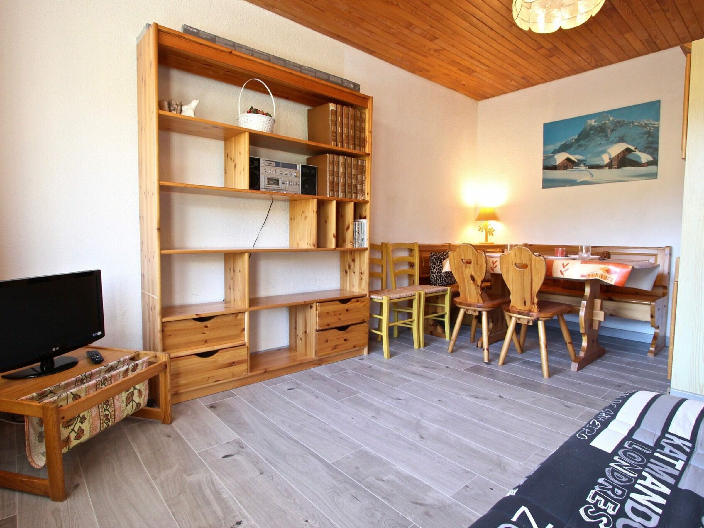 A cozy room with a wooden bookshelf, a desk with a computer, a TV, and a dining area with chairs and a table.