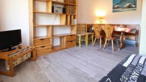 Quarto acolhedor com estante de madeira, escrivaninha com computador, televisão e área de refeições com cadeiras e mesa.