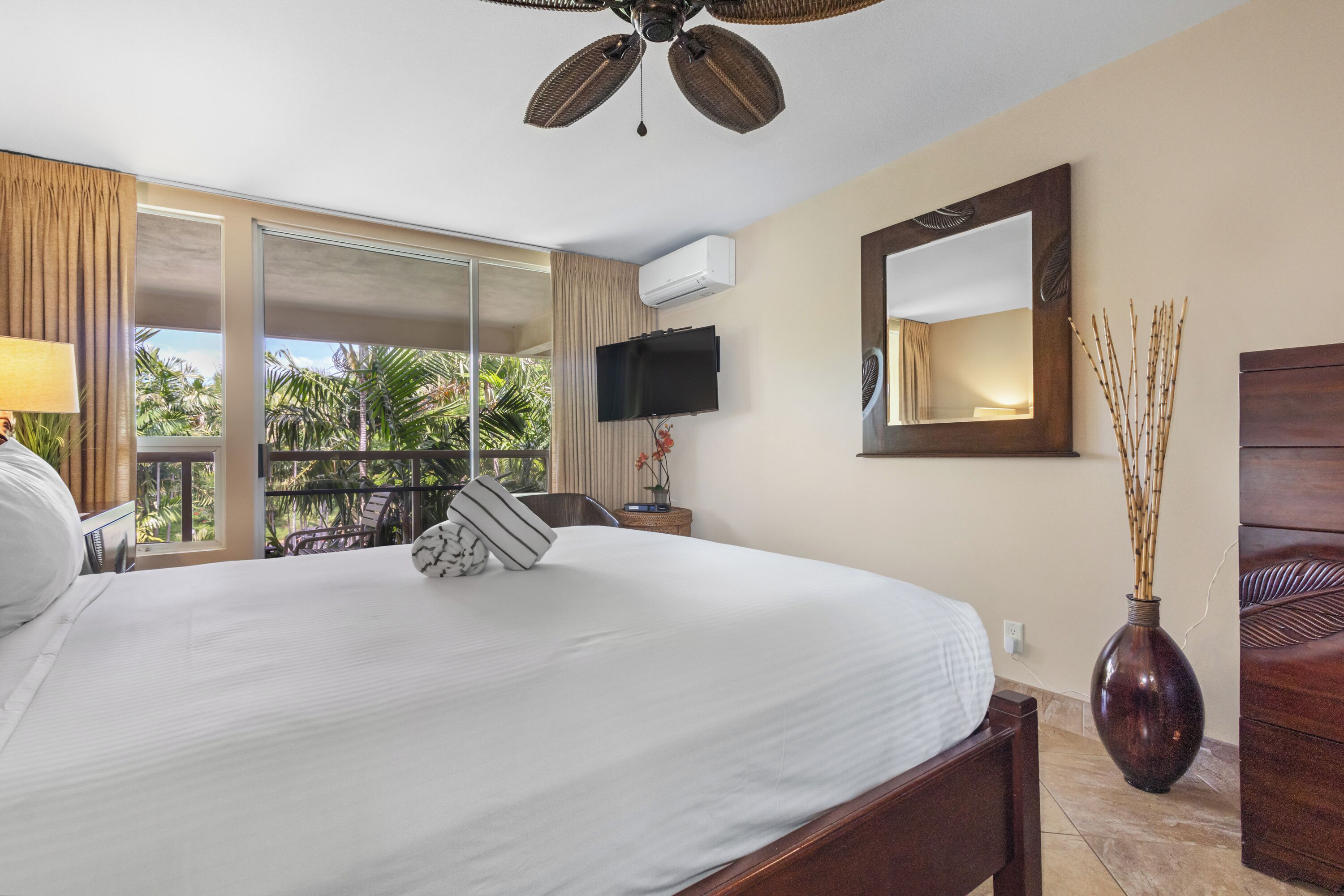 Habitación de hotel con una cama grande, ventilador de techo, televisión, vista a palmeras y puerta corrediza al balcón.