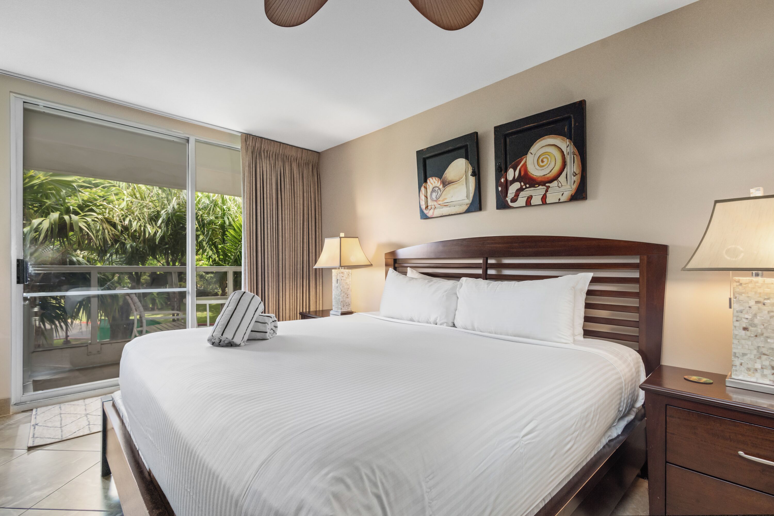 Habitación de hotel con una cama grande, dos obras de arte colgadas en la pared, una mesita de noche con lámpara y vistas a zonas verdes a través de una puerta corrediza.