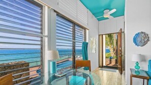 A room with a glass table, chairs, a view of the ocean, a ceiling fan, and a door leading outside.