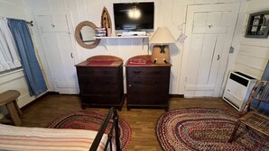 A cozy room with a wooden ceiling, a television on a shelf, two dark wooden dressers, a lamp, and two patterned rugs on the floor.