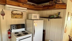 A compact kitchen with a white stove, a white refrigerator, and a microwave on top. There is a wooden shelf above the refrigerator and a fire extinguisher mounted on the wall.