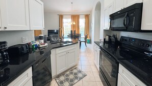 Una cocina moderna con armarios blancos, encimeras negras, microondas integrado y estufa. Se ve un comedor con mesa y sillas a través de una ventana con cortinas.