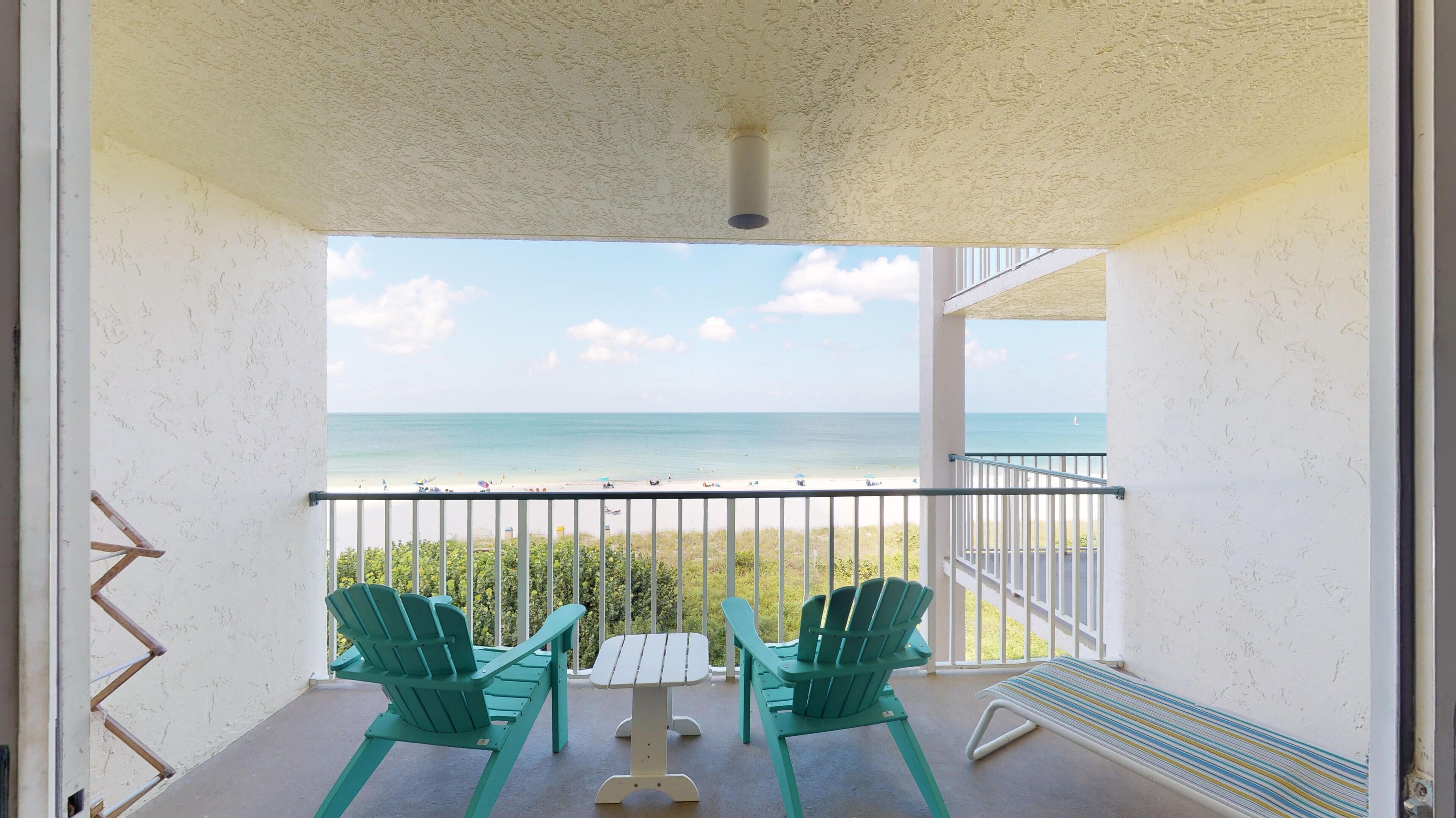 Un balcon avec deux fauteuils Adirondack verts, une petite table et un banc. Le balcon donne sur une plage et offre une vue dégagée sur l’océan.