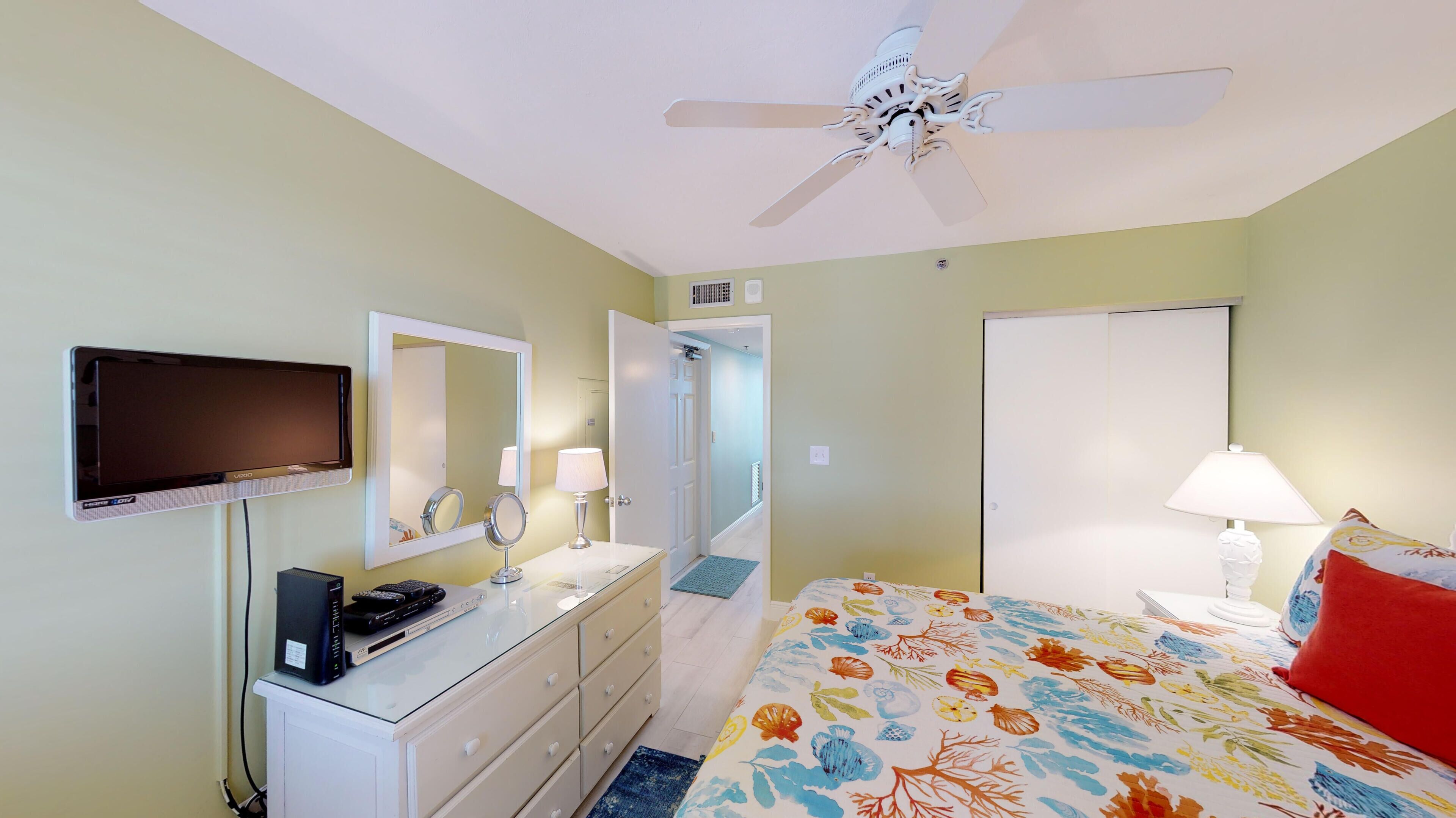 A hotel room with a bed, a TV, a desk with a computer, a mirror, a ceiling fan, and a door leading to another room.