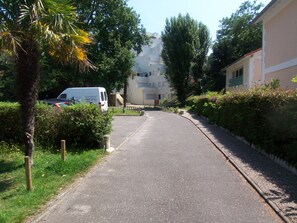 A narrow residential street with a white van parked on the side, surrounded by greenery and a building with a white facade.