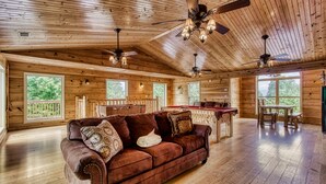 A spacious wooden interior with a large brown leather sofa, ceiling fans, and large windows offering views of greenery. - Big Bear Lodge: Mountain Views, Game Room (Sevierville)