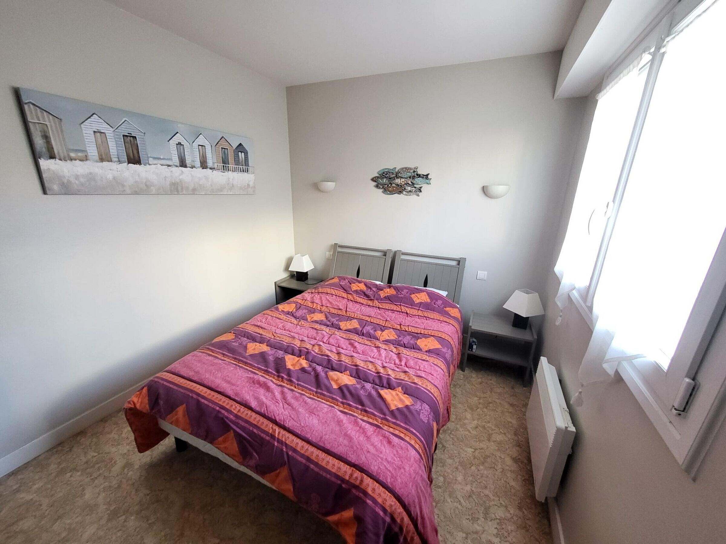 A bedroom with a bed, a nightstand, a wall-mounted lamp, a radiator, and a window.