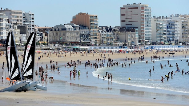 A beach with people, a sailboat, and buildings in the background. - 2-Bedroom Apartment Sea View - Parking, Pets Allowed (Les Sables-d'Olonne)