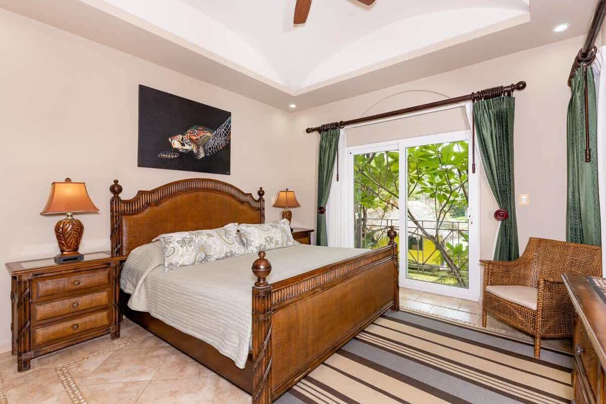 A bedroom with a wooden bed, two bedside tables with lamps, a wicker chair, and a view through a glass door to the outside.