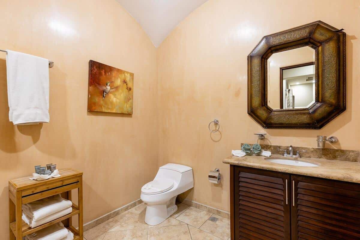 Un baño con un inodoro blanco, un tocador de madera con un lavamanos, un perchero y un cuadro en la pared.