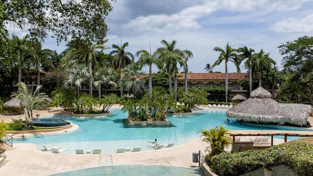 Ein Poolbereich eines Resorts mit Palmen, einer Struktur mit Reetdach und Liegestühlen.
