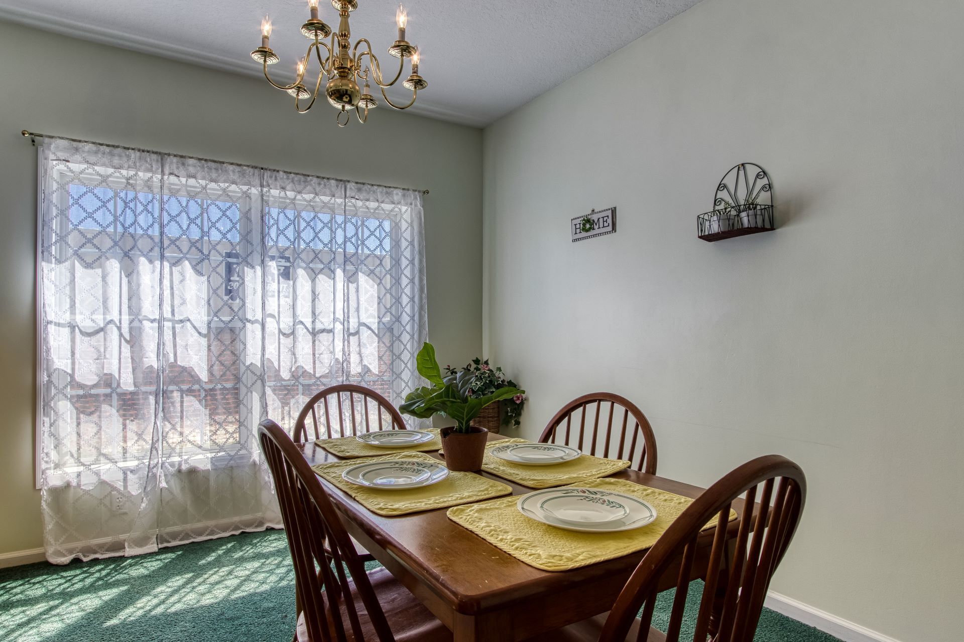 Une salle à manger avec une table dressée pour quatre personnes, un lustre, des rideaux en dentelle et un panneau décoratif représentant le mot « MAISON » accroché au mur.