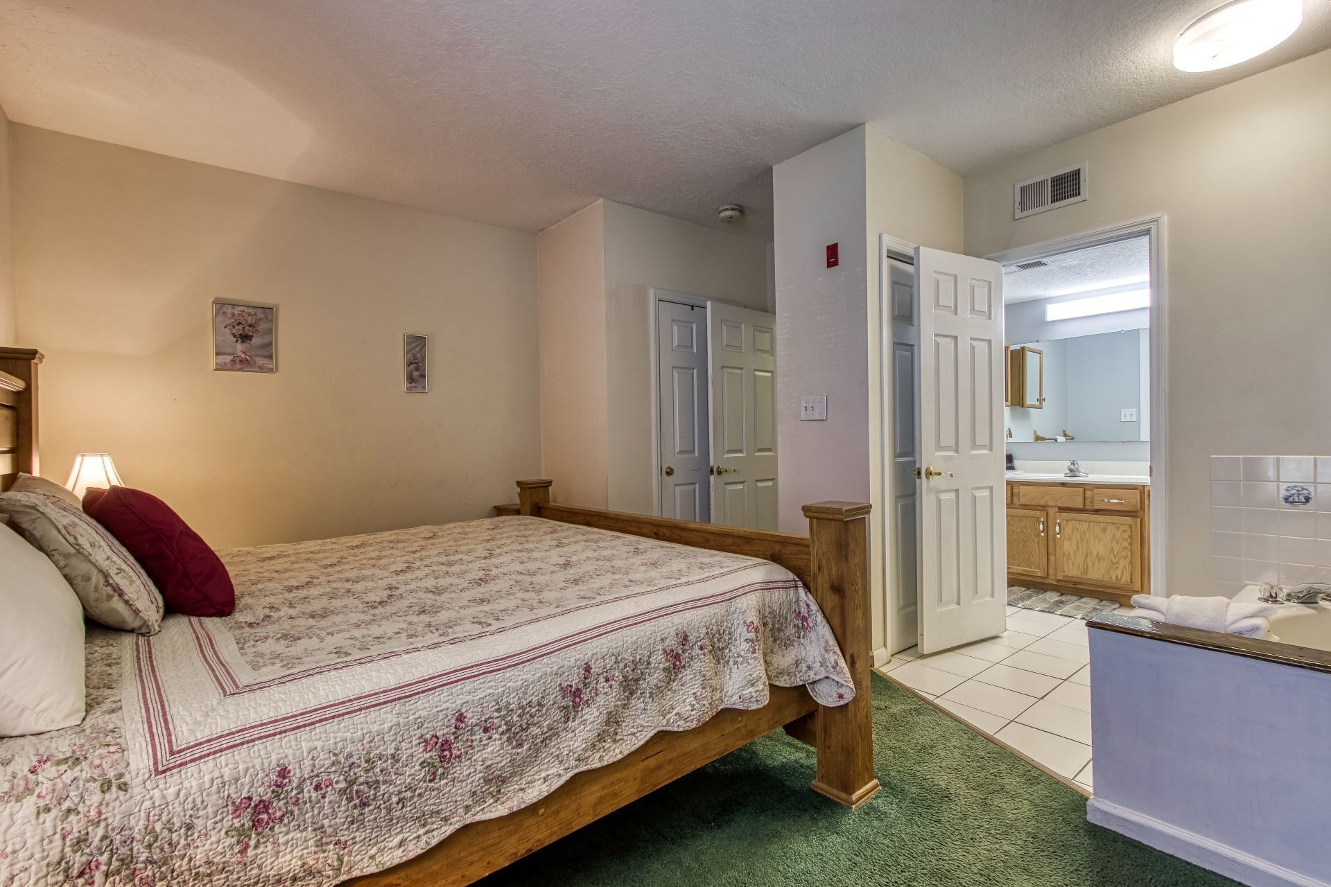 A bedroom with a bed, a nightstand, a lamp, and a door leading to a bathroom with a sink and mirror.