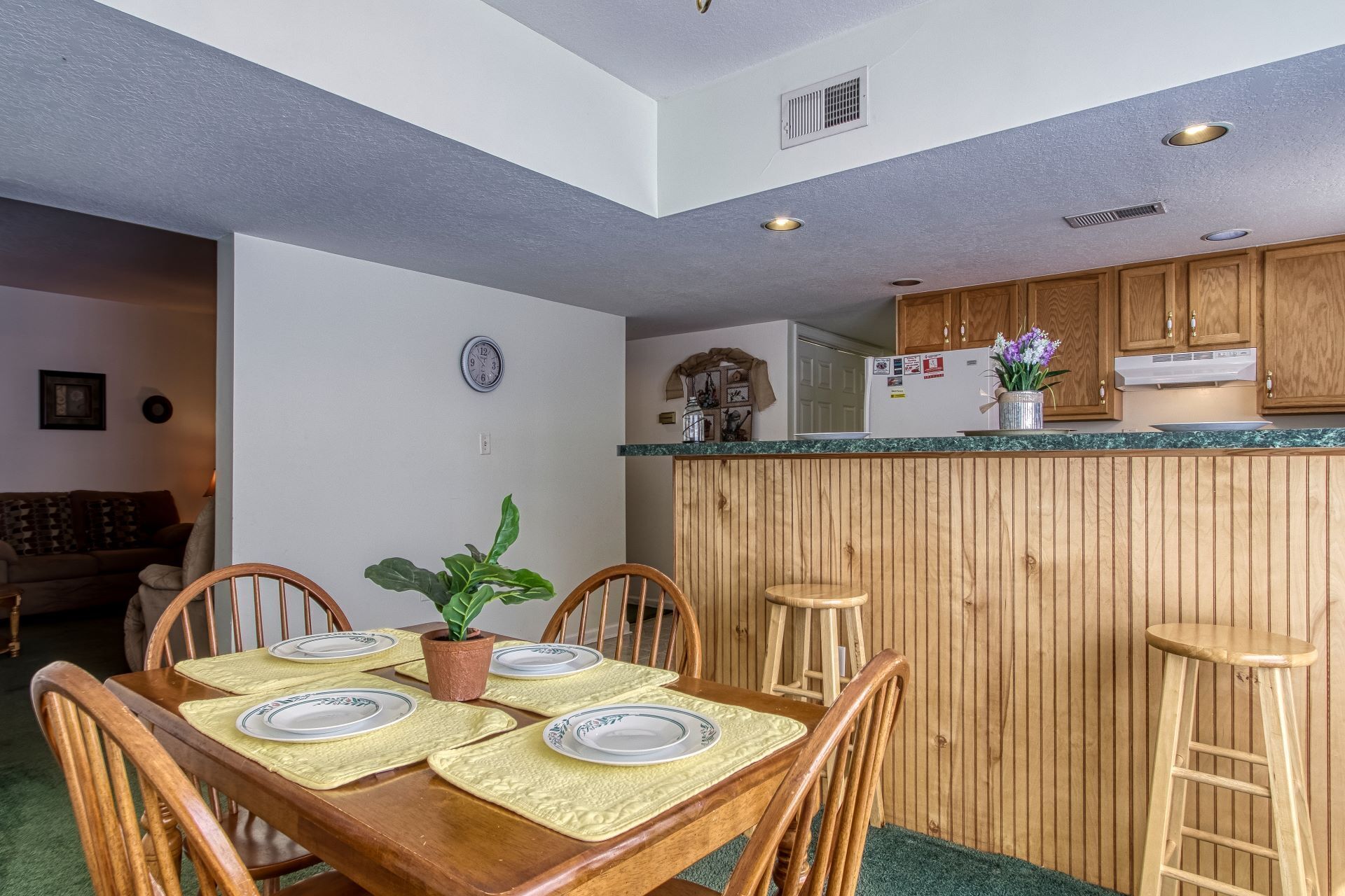 A dining area with a wooden table set for four, a potted plant, and a kitchen in the background.
