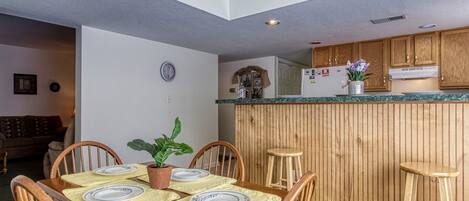 A dining area with a wooden table set for four, a potted plant, and a kitchen in the background.