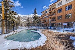 Un espace piscine avec un jacuzzi, entouré d’un chemin pavé en briques et d’un sol recouvert de neige, avec une chaîne de montagnes en arrière-plan et un bâtiment doté de balcons.