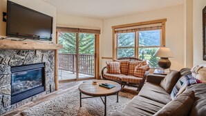 Ein Wohnzimmer mit Kamin aus Stein, einem Flachbildfernseher, einer Couch, einem Couchtisch und Blick auf eine Terrasse mit Holzgeländer.