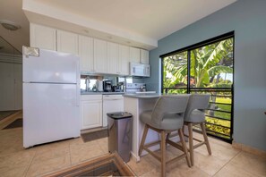 A compact kitchen with white cabinets, a built-in microwave, and a refrigerator. There is a small trash bin and a window with a view of greenery. Two chairs are placed near the window.