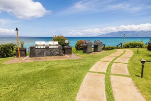A grassy area with barbecue grills and trash bins, a stone pathway, and a view of the ocean. - Oceanfront Kuleana 416 - West Maui Top Floor Condo Fabulous Oceanview (Lahaina)