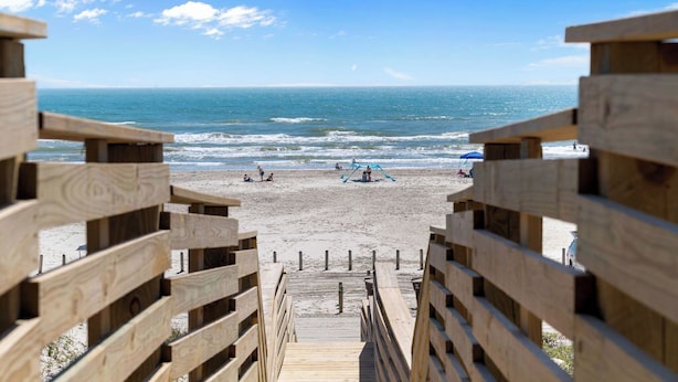 Een houten trap die naar een strand leidt, met een helder uitzicht op de oceaan en mensen in de verte.
