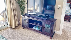 A living room with a dark wooden entertainment center, a large flat-screen TV, a potted plant, and a view of a bedroom with a bed and a chair.