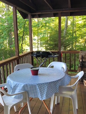 Un espace extérieur abrité, aménagé pour les repas, avec une table ronde et des chaises en plastique blanches, entouré d’une terrasse en bois et offrant une vue sur une forêt.