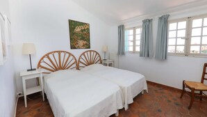 A room with two beds, a small table, a chair, a window with curtains, and a painting on the wall.
